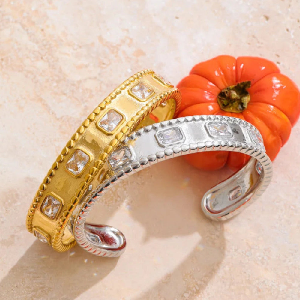 Two gold and silver bracelets on a textured beige surface with a small orange pumpkin. Two gold and silver bracelets on a textured beige surface with a small orange pumpkin.