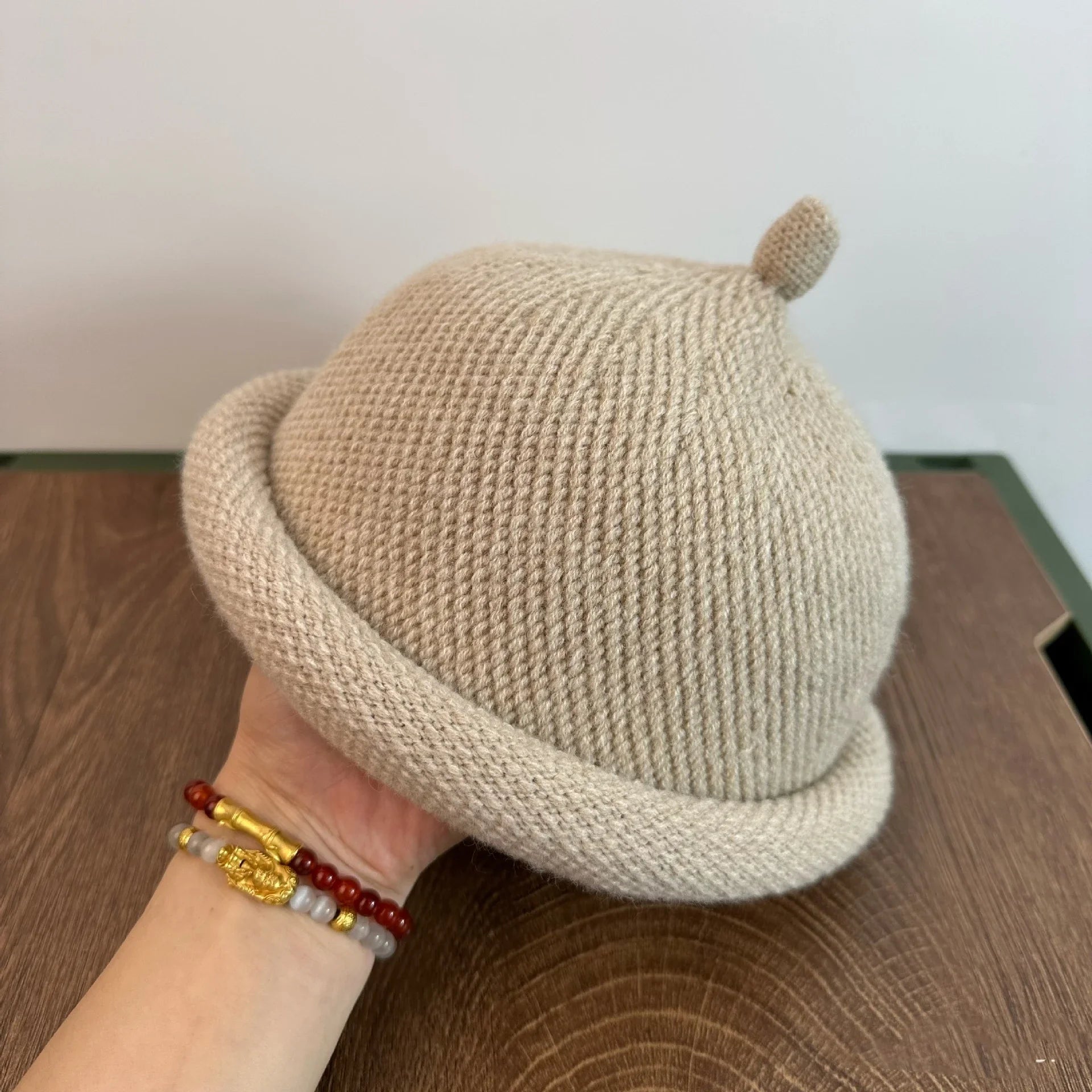 Beige knit hat with a small pom-pom held by a hand on a wooden surface. Beige knit hat with a small pom-pom held by a hand on a wooden surface.